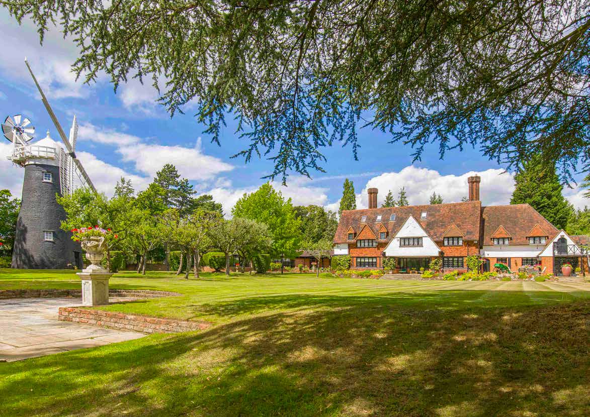 A large red brick country house with tall chimneys seen across a lawn, with a black weatherboarded windmill beside it