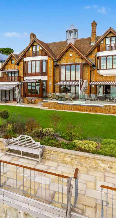 A large red brick and tile hung waterfront house with striped awnings, seen across a lawn and a stone terrace