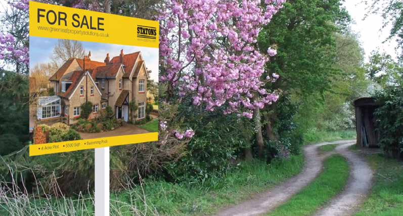 A yellow bespoke for sale board carrying a photograph of a period house, standing at the end of a country lane under blossom