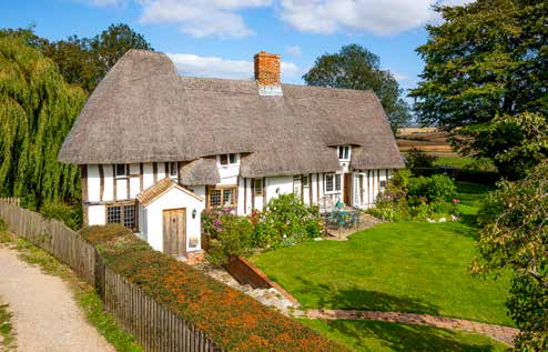 A white and black timbered thatched cottage behind a picket fence with a mown lawn and orange flower beds