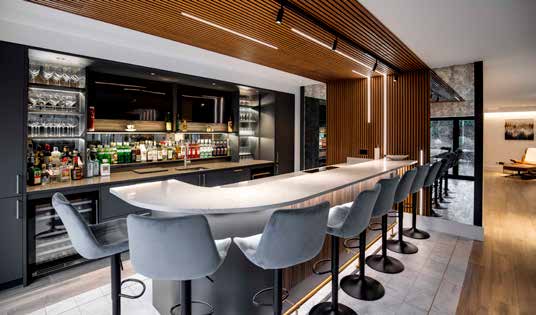 A home bar with a curved white counter, grey velvet stools and a slatted timber ceiling