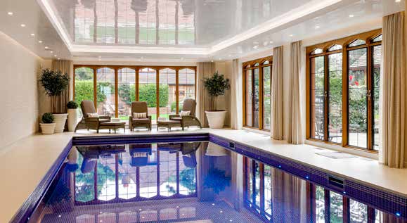 An indoor swimming pool with deep blue tiling, arched windows and wicker chairs along the far end