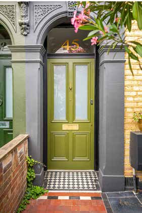 An olive green panelled front door under a stone arch, with a black and white tiled step and pink oleander overhead