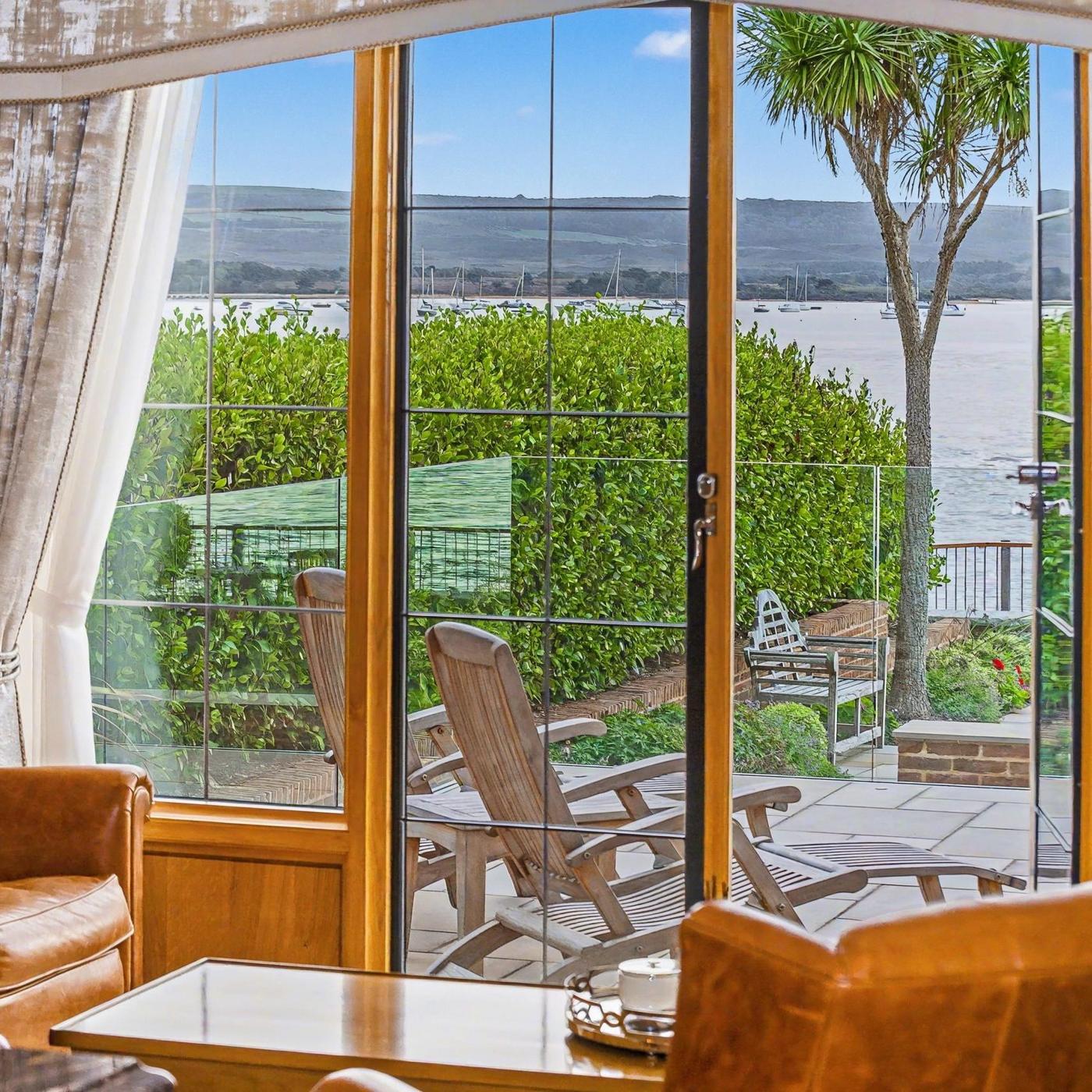 A sitting room with leather armchairs looking through tall glass doors to a terrace, a harbour and moored yachts