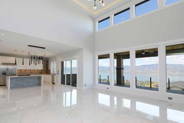An empty open plan living space with a polished marble floor, an open kitchen behind and tall windows looking out over a lake and hills