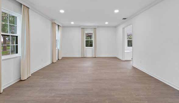 An empty room with a wood floor, white walls, cream curtains at three windows and recessed ceiling lights